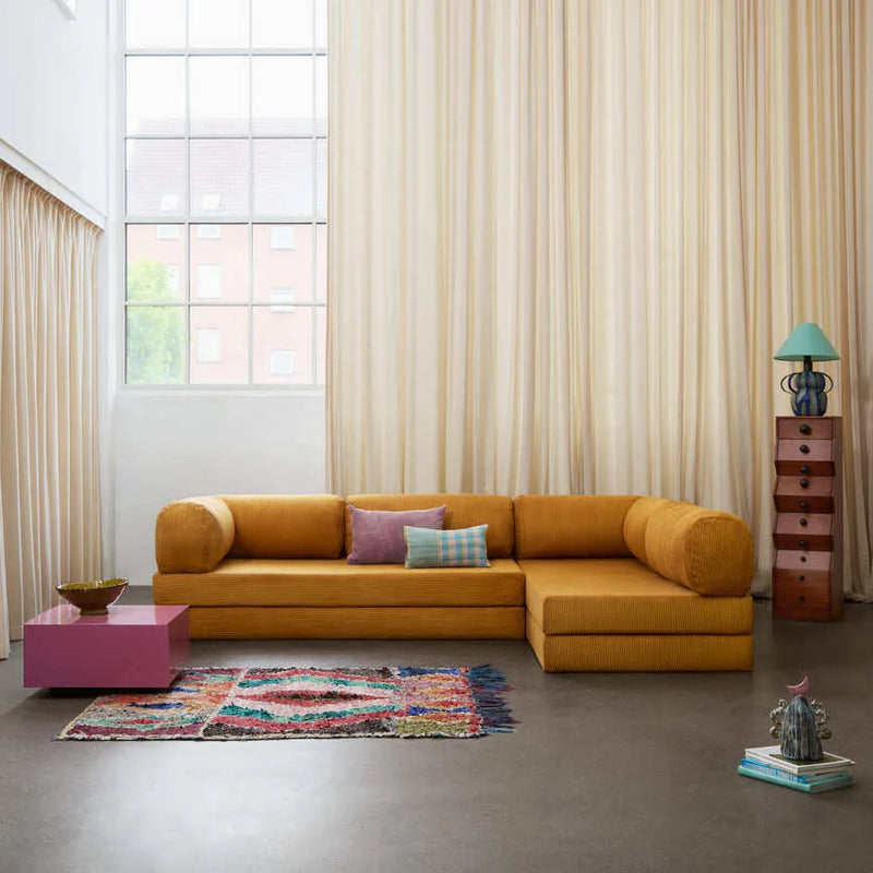 A modern mustard yellow corduroy l-shaped sectional sofa with cylindrical arms is styled in a bright, minimalist Scandinavian living room with floor-to-ceiling cream curtains and colorful geometric rug.